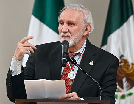 A man in a suit and tie speaks into a microphone at a podium with Mexican flags behind him.